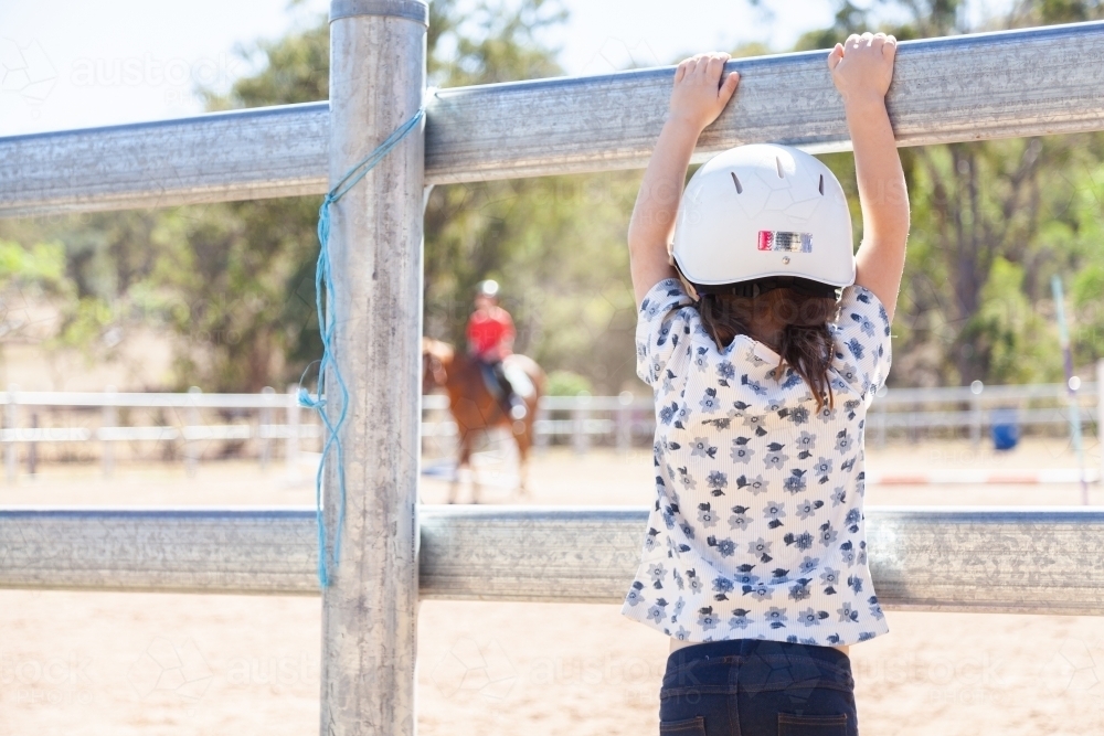 Little girl climbing on arena fence watching riding lesson - Australian Stock Image