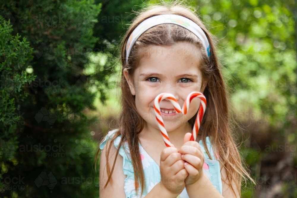 Little girl holding candy cane sweets in heart shape outside near pine tree - Australian Stock Image