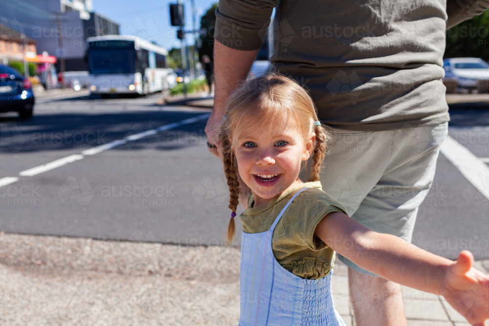 Little girl holding dads hand waiting to cross road at intersection in city with traffic lights - Australian Stock Image