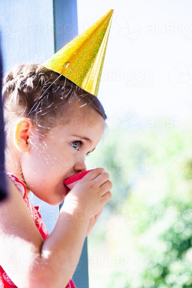 Little girl in party hat eating cupcake - Australian Stock Image