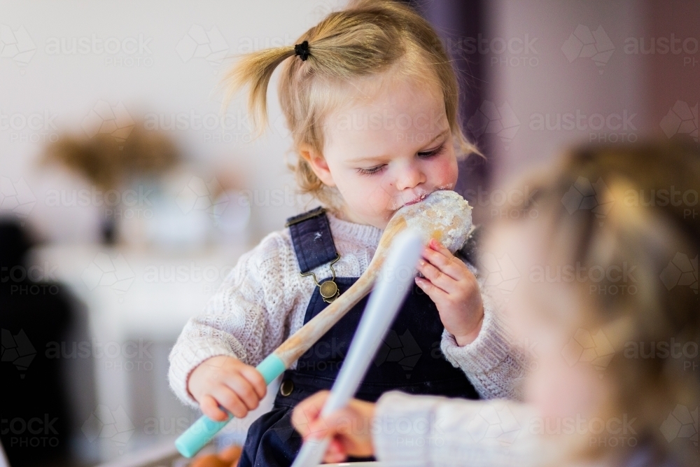 Little girl licking mixing spoon while helping mum bake in kitchen - Australian Stock Image