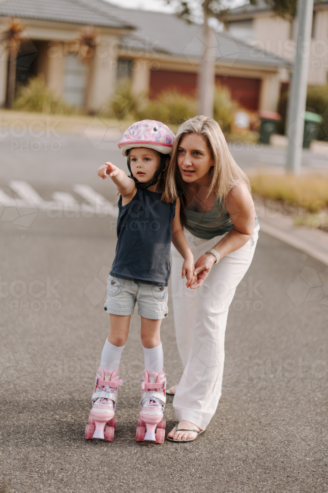 Little girl on roller skates and wearing a pink helmet standing on the street with mum. - Australian Stock Image