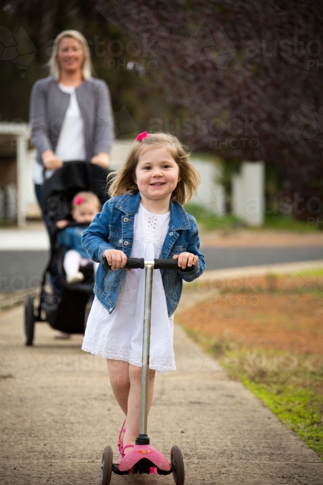 Little girl on scooter on footpath with mother behind - Australian Stock Image
