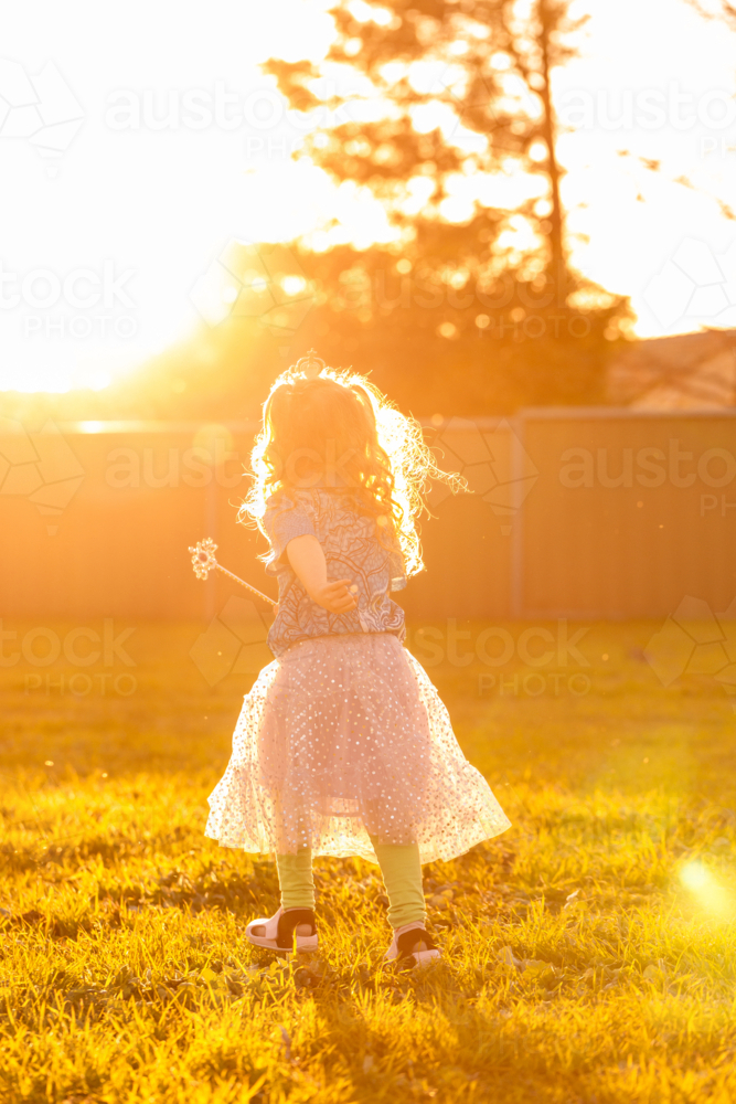 Little girl playing dressups with princess skirt and wand running in sunshine in backyard - Australian Stock Image