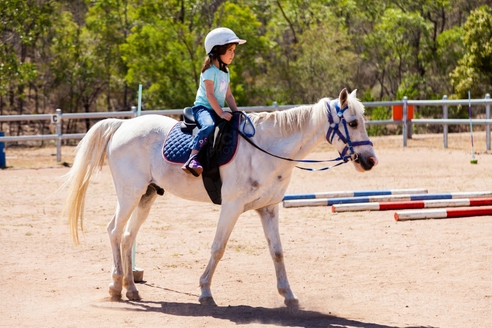 Little girl riding a white pony in dusty yard at riding school - Australian Stock Image