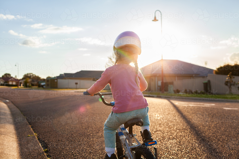 Little girl riding bike down quiet suburban road with backlighting at sunset - Australian Stock Image
