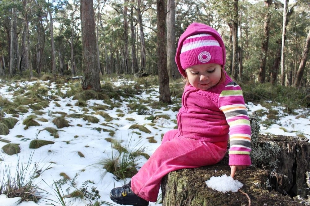 Little girl sitting on a tree stump touching a pile of snow - Australian Stock Image