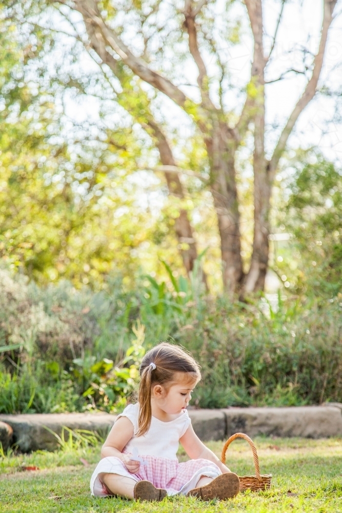 Little girl sitting on green grass in the garden with basket - Australian Stock Image