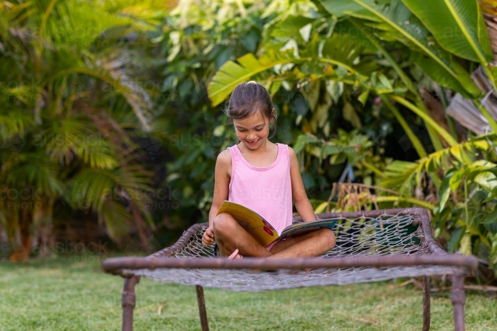 little girl sitting on old bed frame in garden reading a book - Australian Stock Image