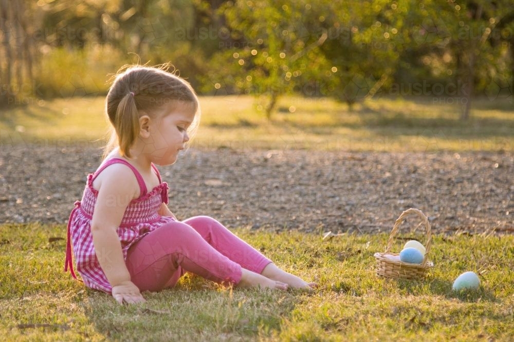 Little girl sitting on the grass near a basket of Easter eggs - Australian Stock Image