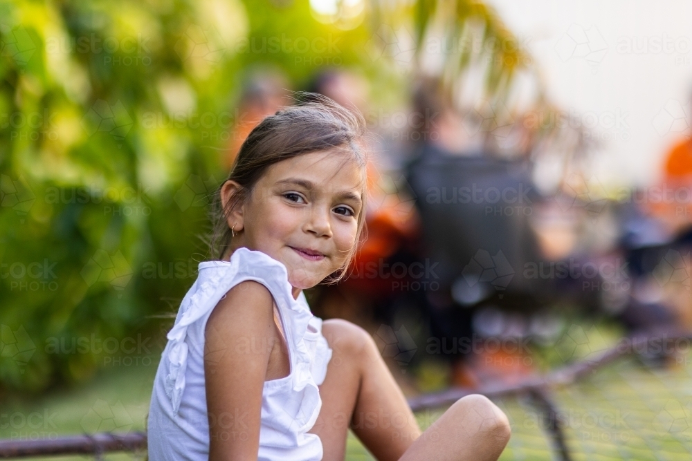 little girl smiling looking at camera over shoulder, with adults blurred in background - Australian Stock Image