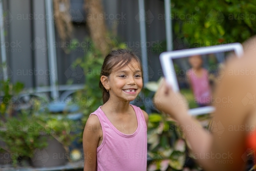 little girl smiling while her mother takes a photo of her on digital tablet - Australian Stock Image