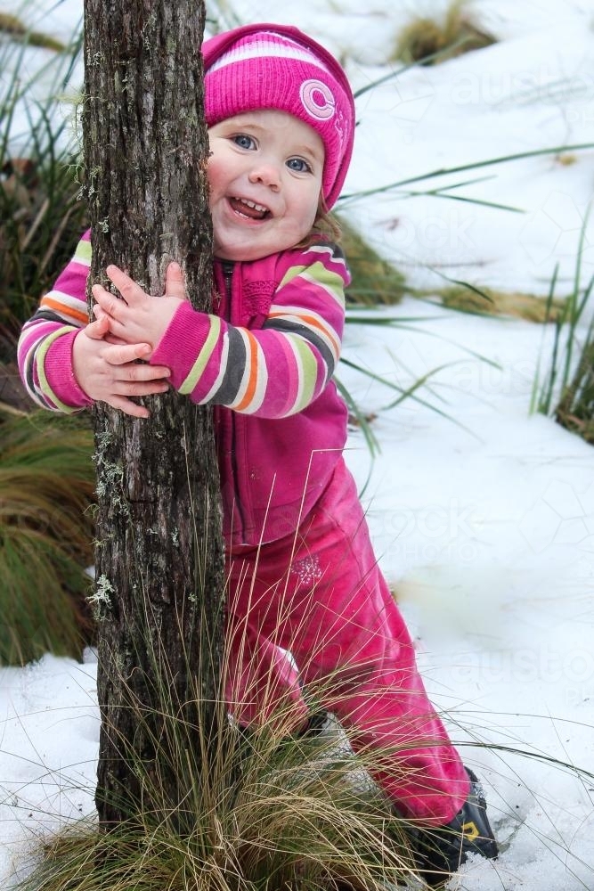 Little girl standing in snow hugging a tree - Australian Stock Image