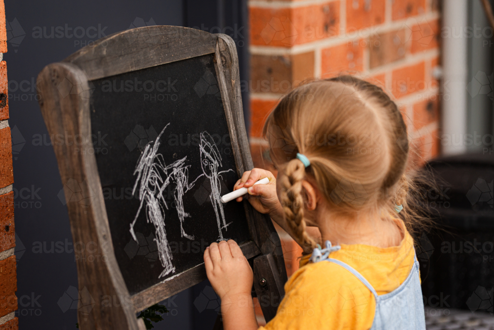 Little girl standing outside in backyard drawing her family on chalkboard - Australian Stock Image