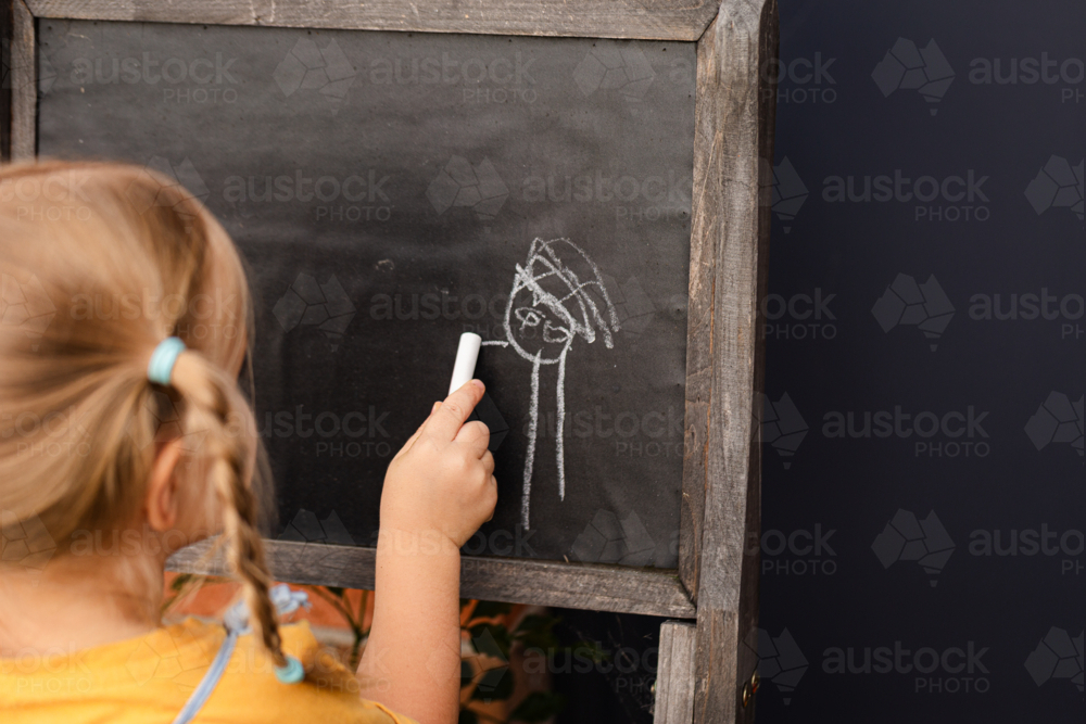Little girl standing outside in backyard drawing person on chalkboard - Australian Stock Image