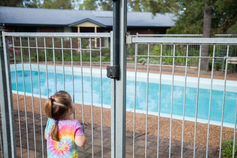 Little girl standing safely outside pool fence gate looking in - Australian Stock Image