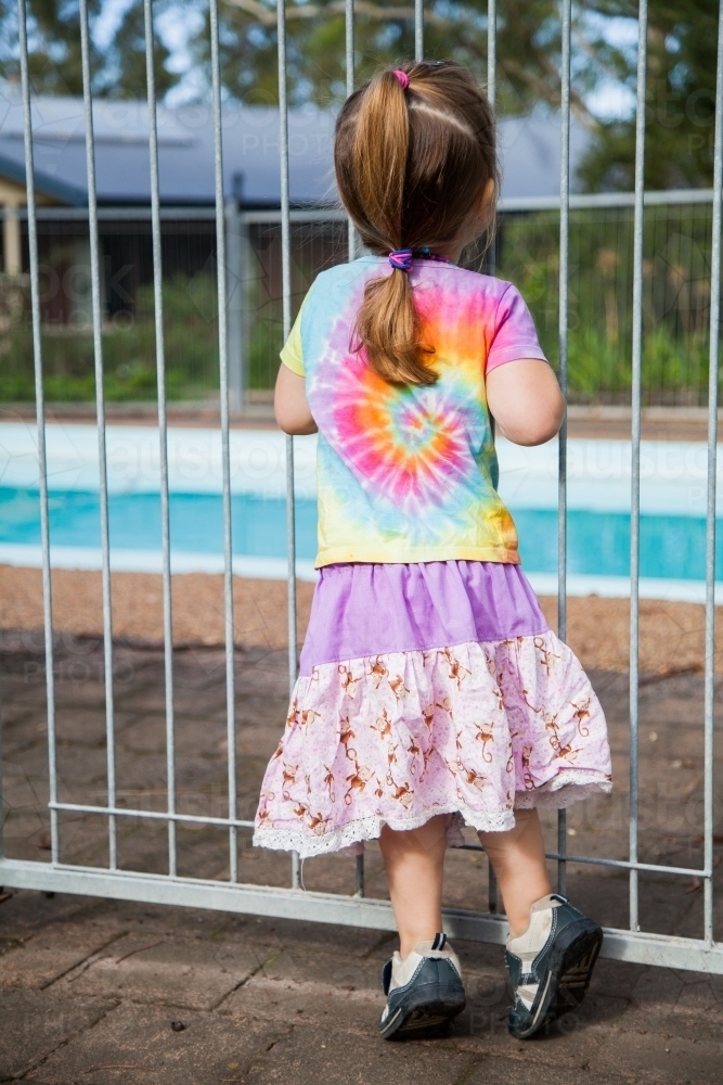 Little girl standing safely outside pool fence gate looking in - Australian Stock Image