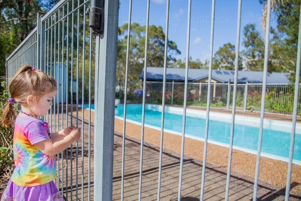 Little girl standing safely outside pool fence gate looking in - Australian Stock Image