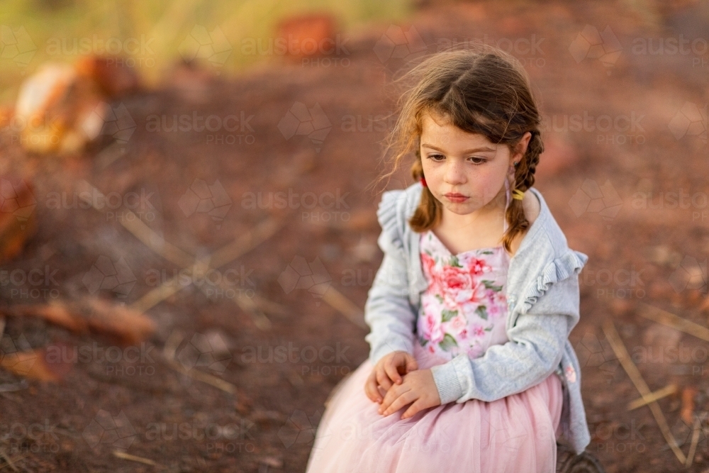 little girl wearing pretty dress sitting by herself outside with bare dirt in background - Australian Stock Image