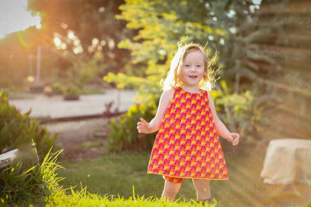 Little girl wearing red dress exploring garden flooded in golden afternoon light - Australian Stock Image