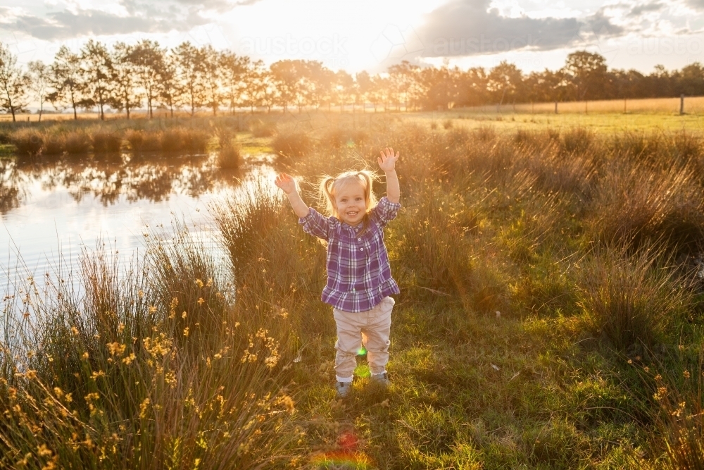 Little girl with arms up saying yay in golden light on farm celebrating after rain - Australian Stock Image