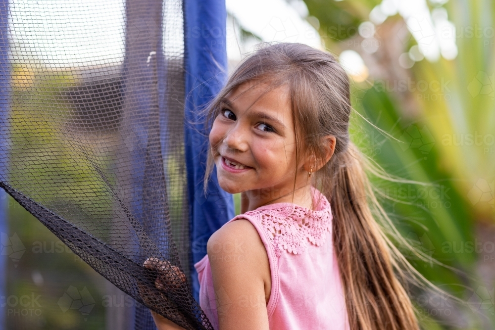 little girl with long hair smiling and glancing over shoulder from the trampoline - Australian Stock Image