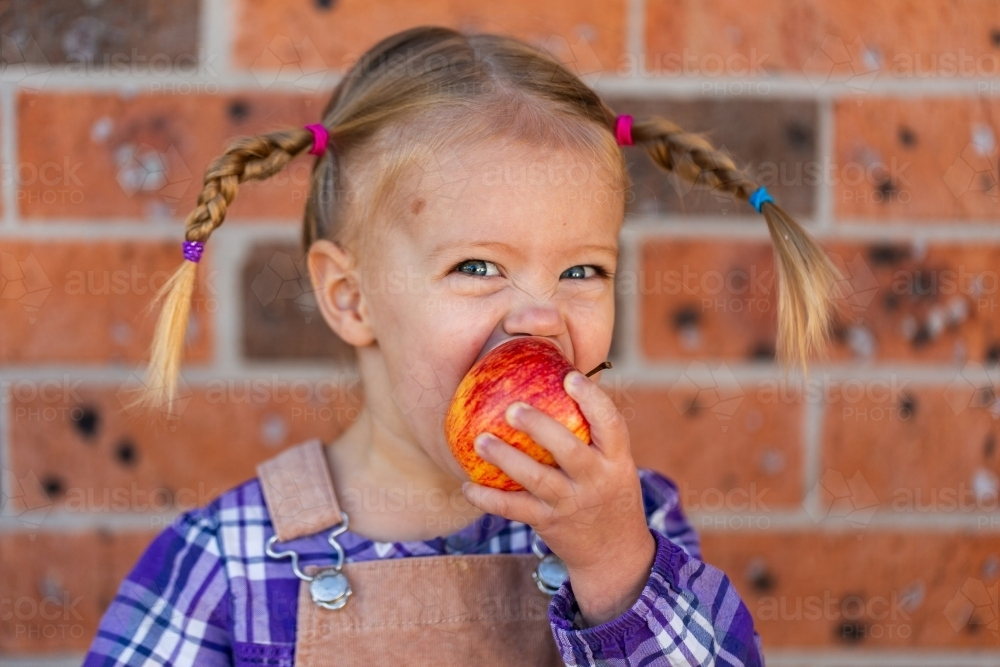 Little kid eating crunchy apple outside by brick wall of home - Australian Stock Image