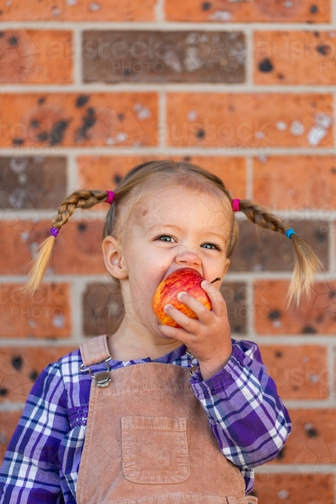 Little kid eating crunchy apple outside by brick wall of home - Australian Stock Image