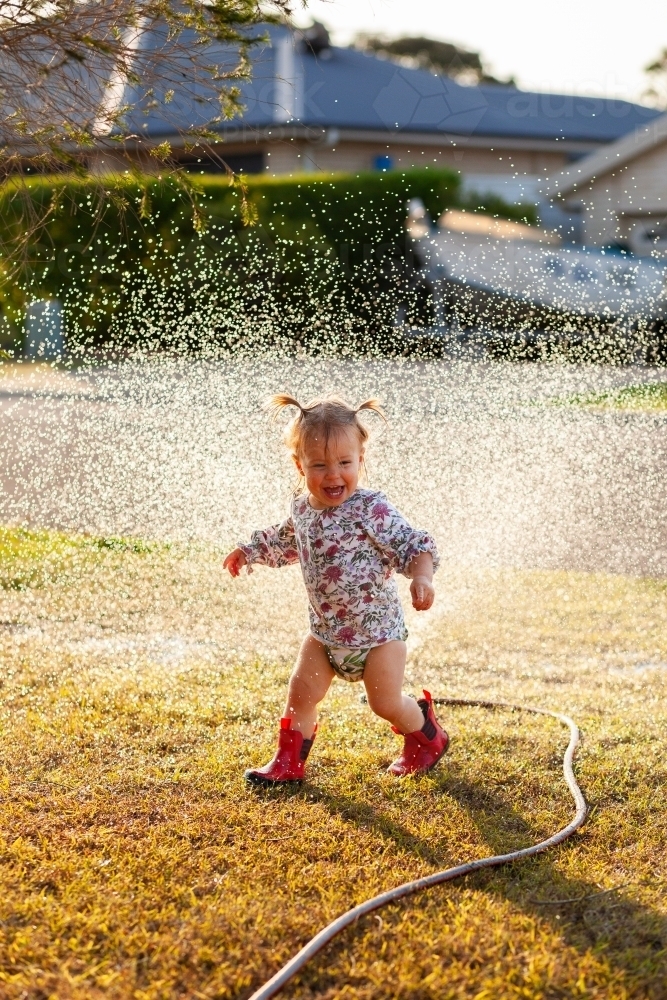 Little kid wearing cloth nappy and red gumboots playing under sprinkler water in yard in summertime - Australian Stock Image