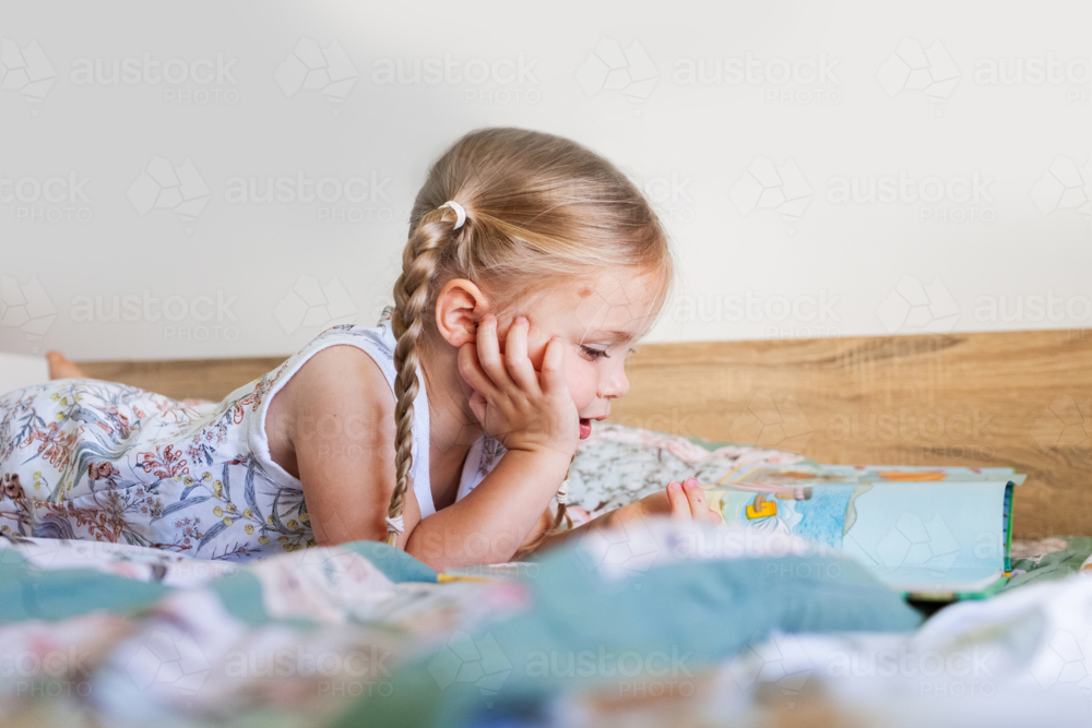 Little preschool aged child lying on bunk bed reading a storybook looking at pictures in bedroom - Australian Stock Image