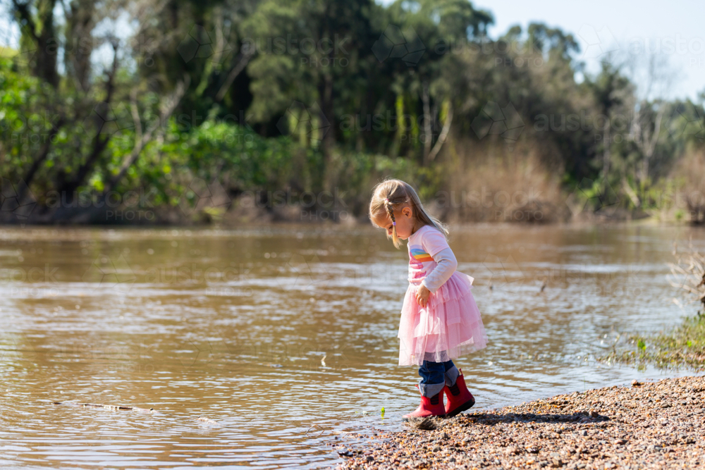Little toddler girl in pink dress and red gumboots playing by riverside - Australian Stock Image