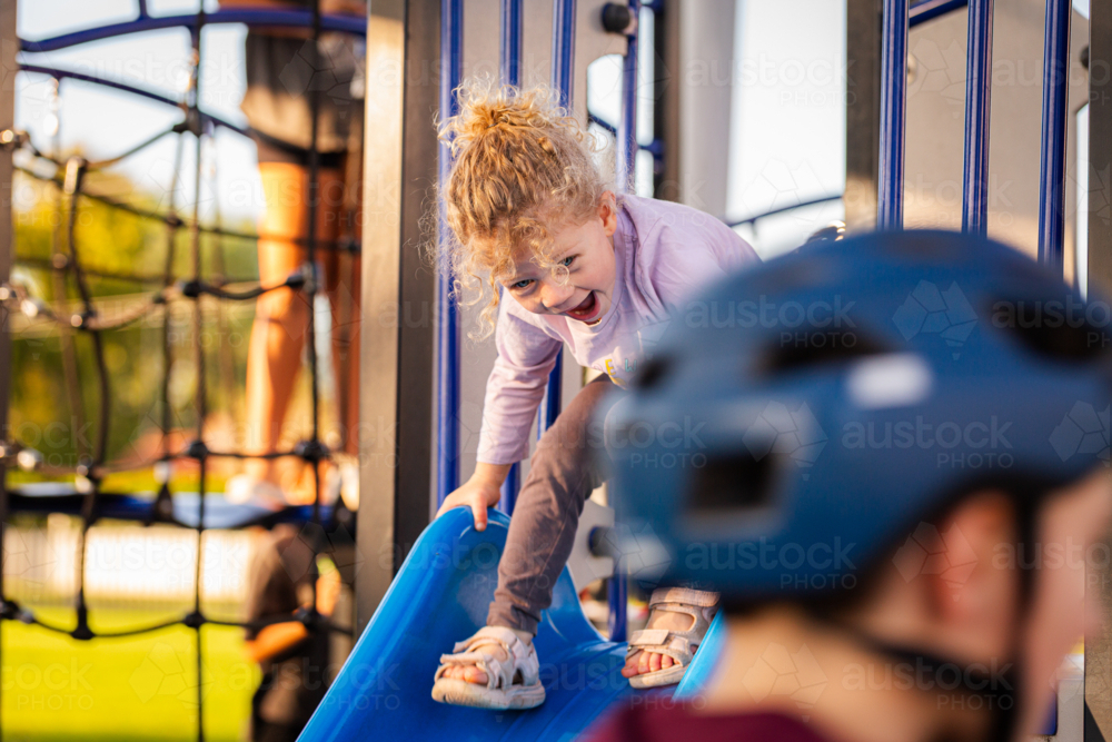 little toddler girl with curly hair playing on slide at park playground - Australian Stock Image