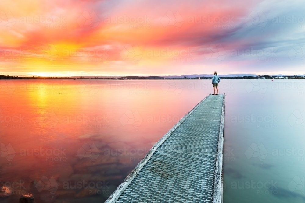 Lone figure standing on a jetty looking out at a vivid sunset - Australian Stock Image