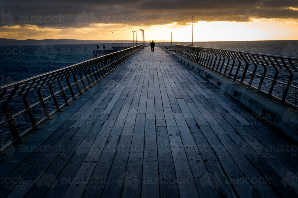 Lone figure walking a weathered pier toward the rising light. - Australian Stock Image
