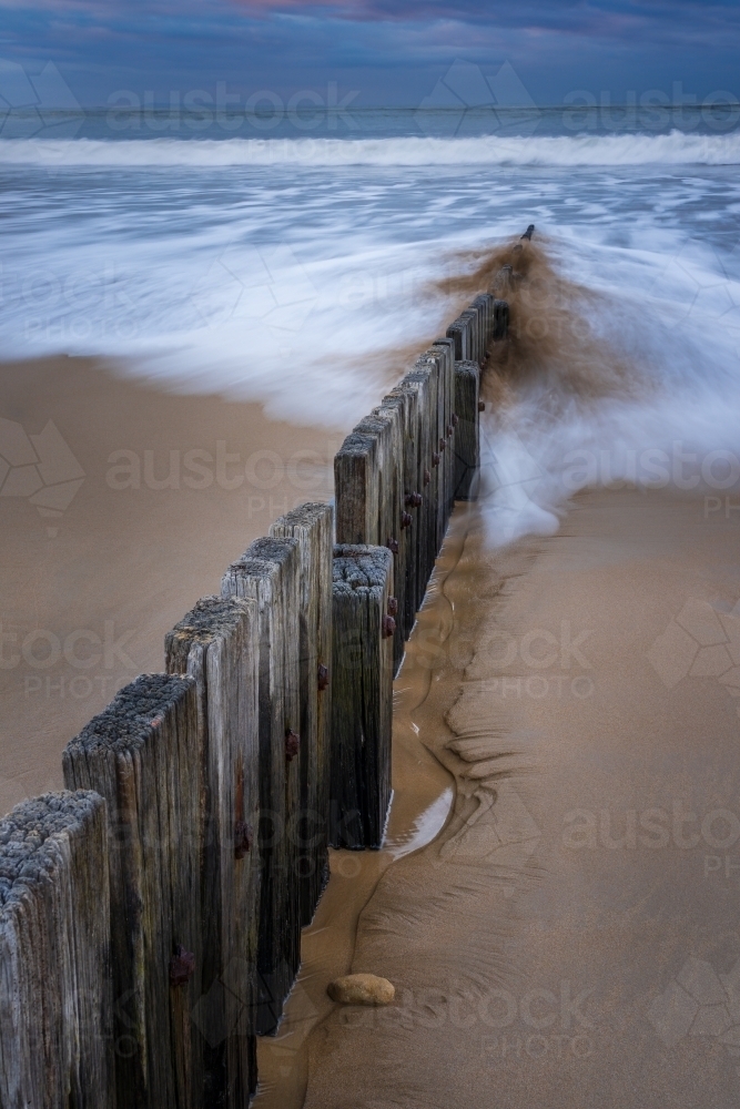 Long exposure of waves crashing over a wooden groyne on a sand beach - Australian Stock Image