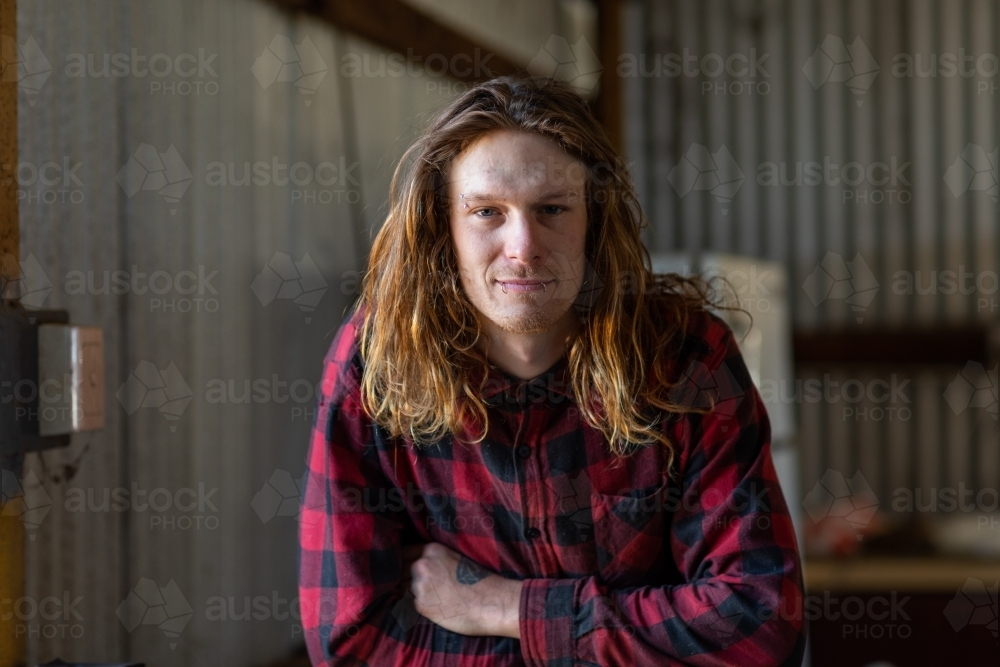 Long-haired guy in red-checked shirt with arms crossed in industrial setting - Australian Stock Image