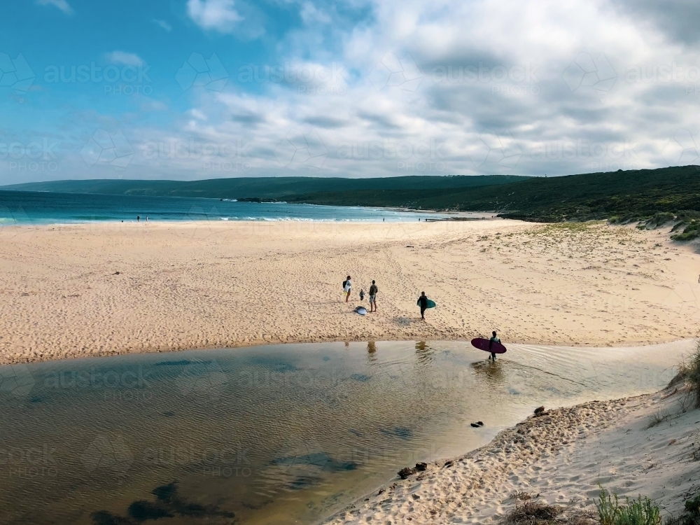Long shot of coastal scene with people crossing river heading to beach carrying surfboards - Australian Stock Image