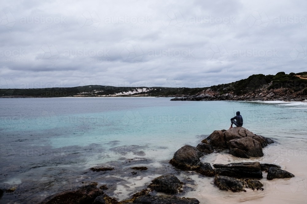 Long shot of man sitting and fishing from rock on remote beach - Australian Stock Image