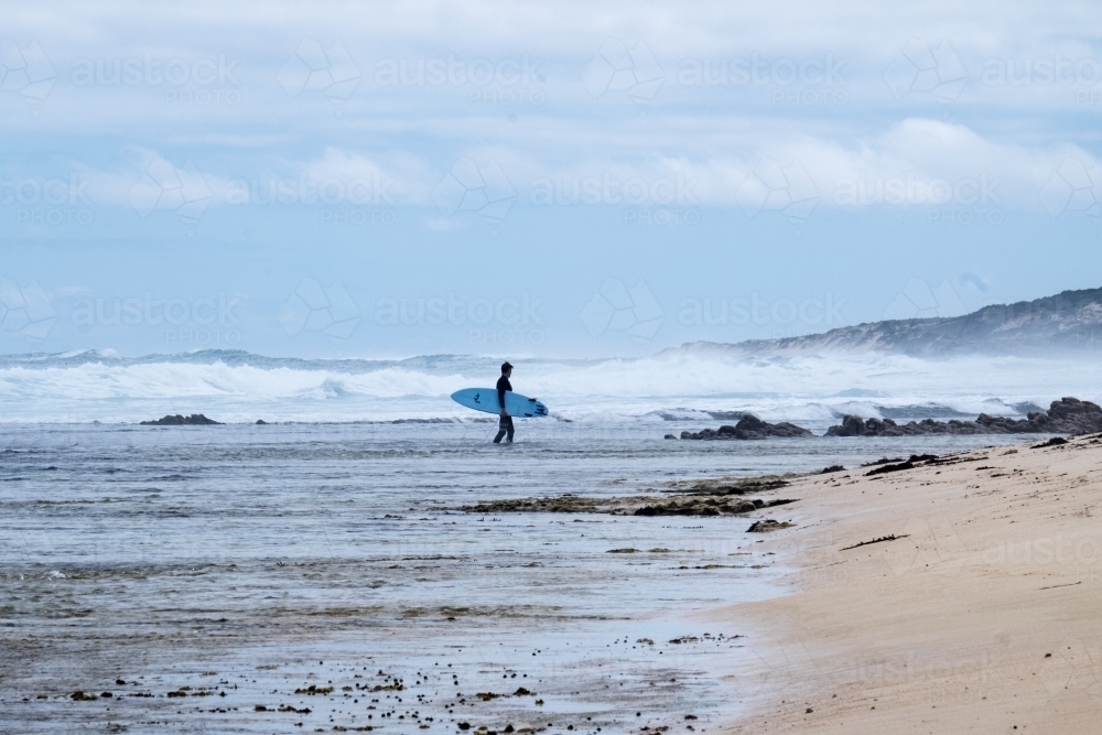 Long shot of Surfer walking in from surf over reef in remote rugged surfing location - Australian Stock Image