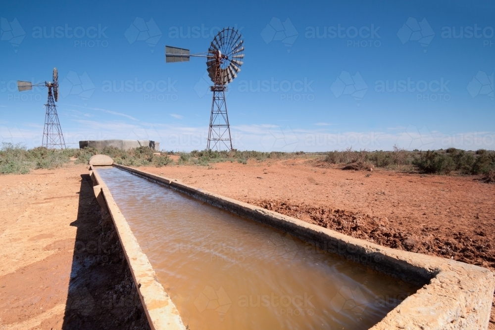 Looking along a full water trough with windmills and tanks in the background - Australian Stock Image