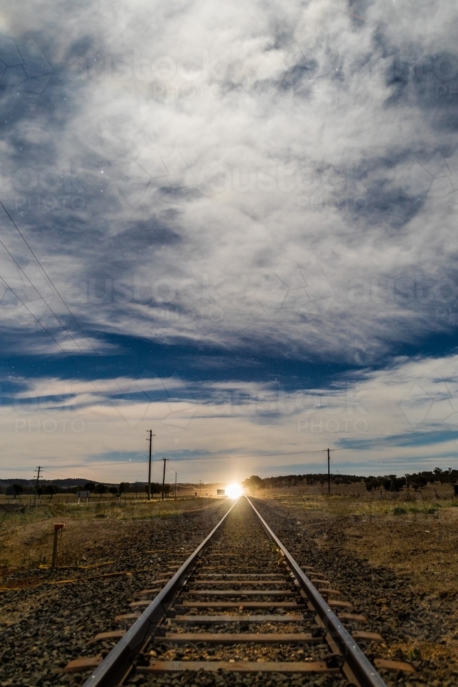 Looking down a Railway track with shining light in the distance on a starry cloudy night - Australian Stock Image