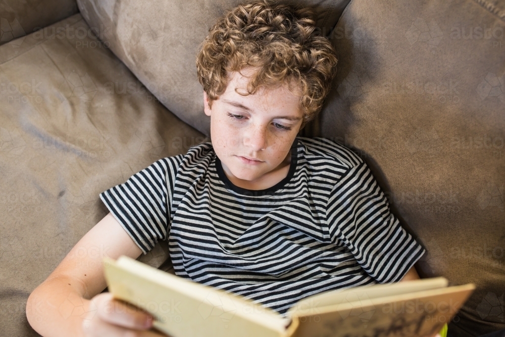Looking down at boy lying on lounge reading book - Australian Stock Image