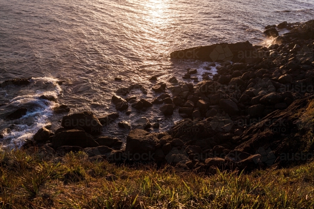 Looking down from a grassy headland as ocean waves crash against large rocks at sunset. - Australian Stock Image