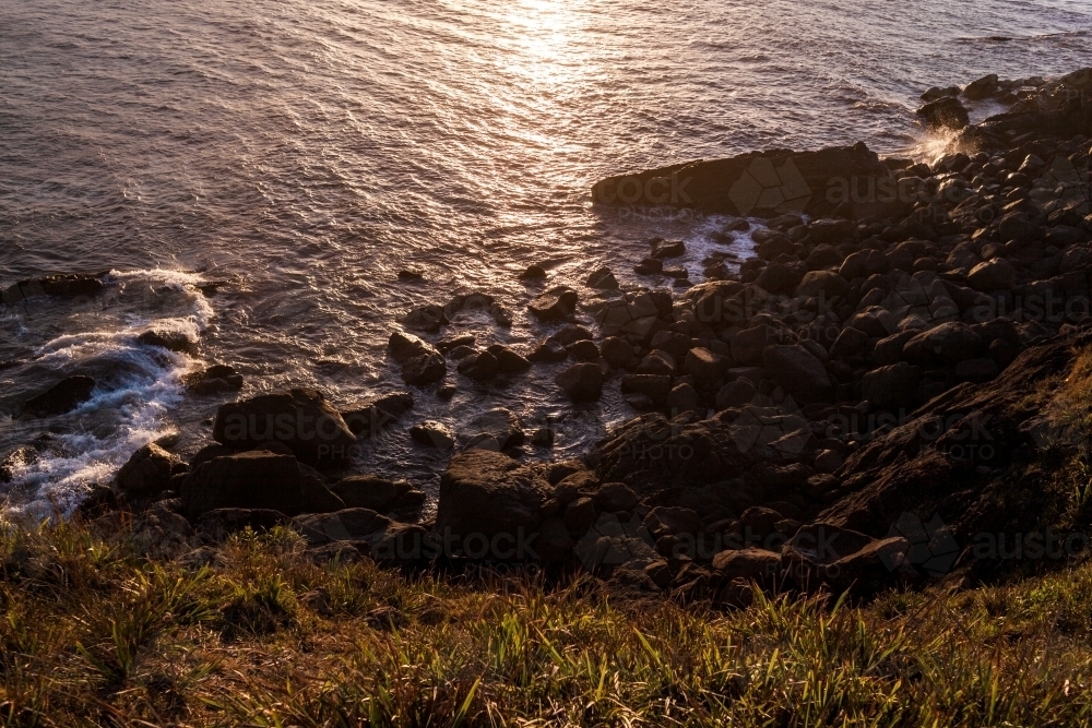 Looking down from a grassy headland as ocean waves crash against large rocks at sunset. - Australian Stock Image