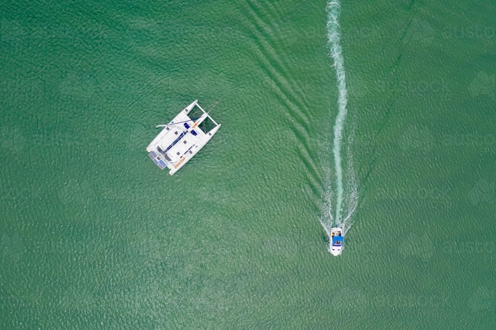 Looking down on a single catamaran and a small boat speeding past leaving a wake in the calm water - Australian Stock Image