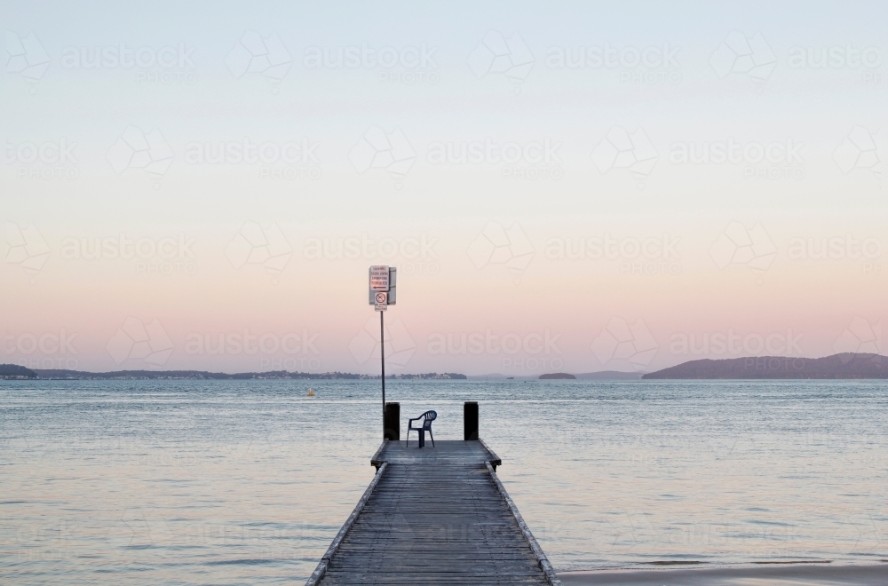 Looking out along a pier at dusk with a single chair at the end - Australian Stock Image