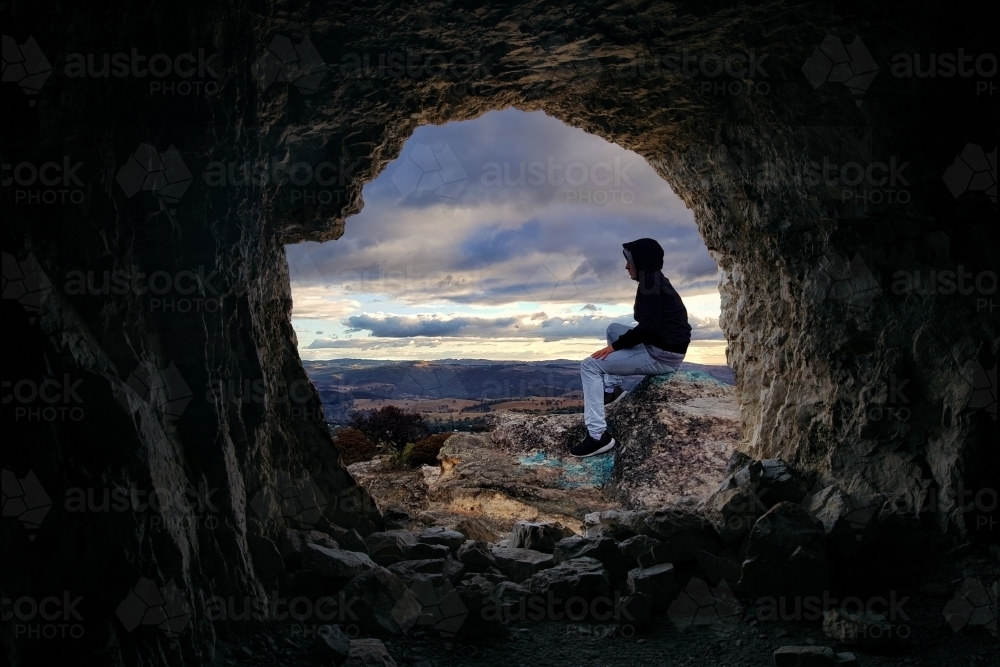 Image of Looking through a cave to a male sitting on a rocky outcrop ...