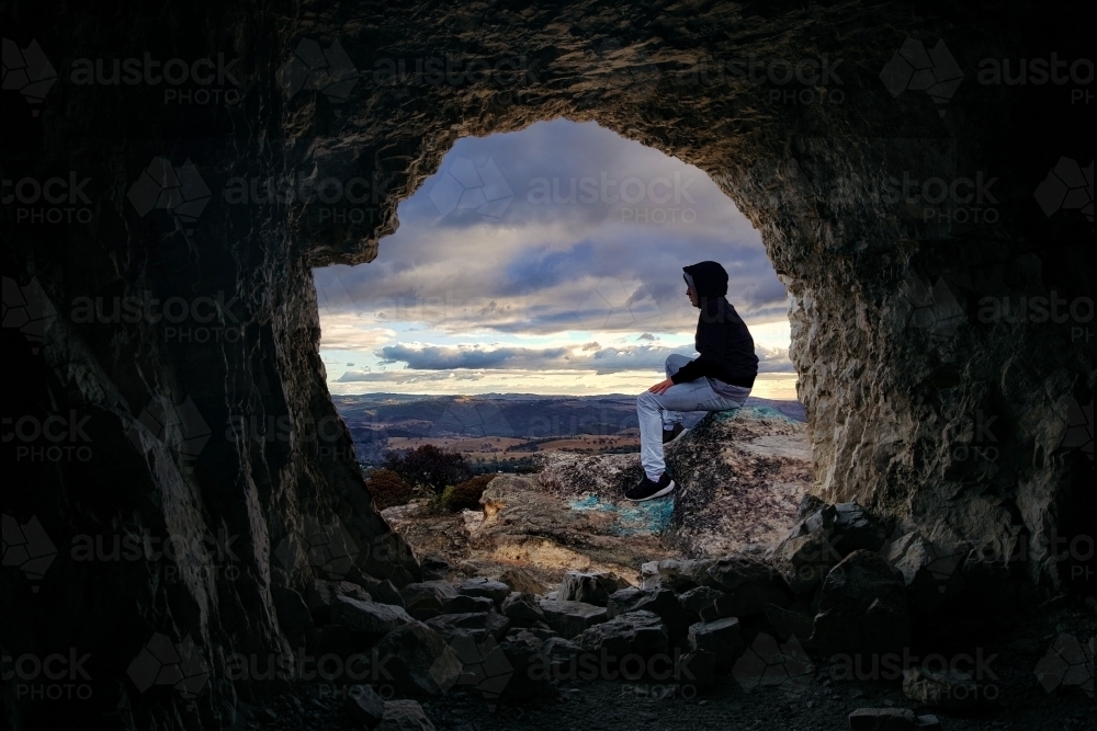 Looking through a cave to a male sitting on a rocky outcrop that looks over the valley - Australian Stock Image