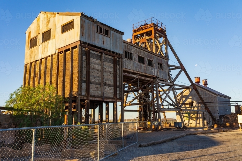 Looking up at the abandoned ruins of a mining site with large shed and poppet head tower - Australian Stock Image