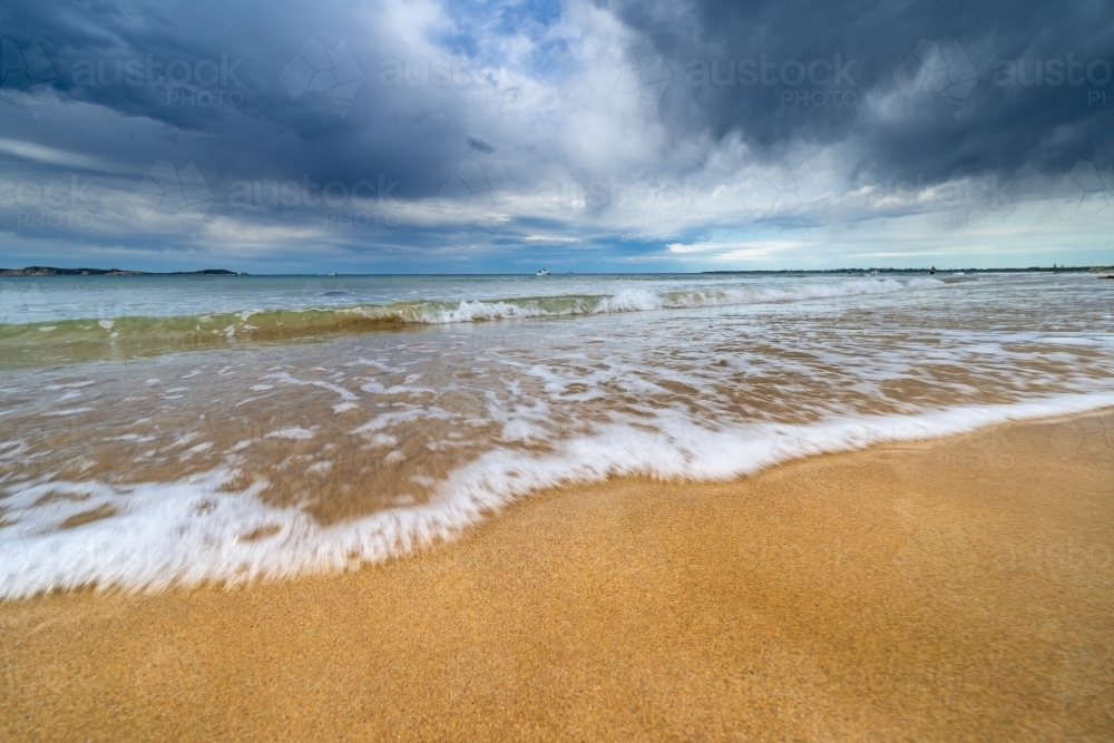 Low angle view of gentle waves rolling up a golden sandy beach with dark storm clouds overhead - Australian Stock Image